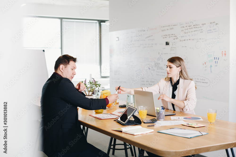 Authentic image of businessman and businesswoman sitting at table and signing finalized documents in modern open office. Authentic image of businessman and businesswoman sitting at table and signing finalized documents in modern open office.