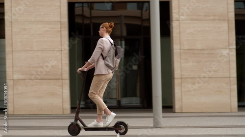 Following shot of young Caucasian woman with red hair wearing eyeglasses riding electric kick scooter in city center