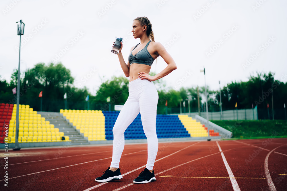 Fototapeta premium Thoughtful adult athletic female holding water bottle in stadium