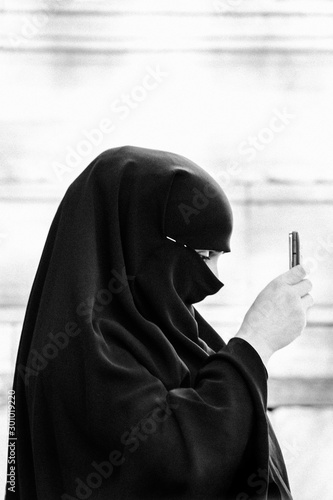 black and white shot of a muslim woman with phone viewed from the side with her face covered by the veil