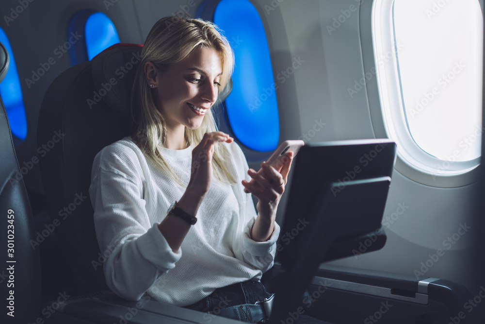 Smiling caucasian female passenger texting message on mobile phone while enjoying comfortable flight with Wireless network access on board. Young tourist woman choose airline with internet connection Smiling caucasian female passenger texting message on mobile phone while enjoying comfortable flight with Wireless network access on board. Young tourist woman choose airline with internet connection