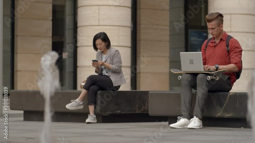 Lockdown of young Caucasian man with skateboard and laptop on his knees and Asian young woman with smartphone in her hands sitting in city center