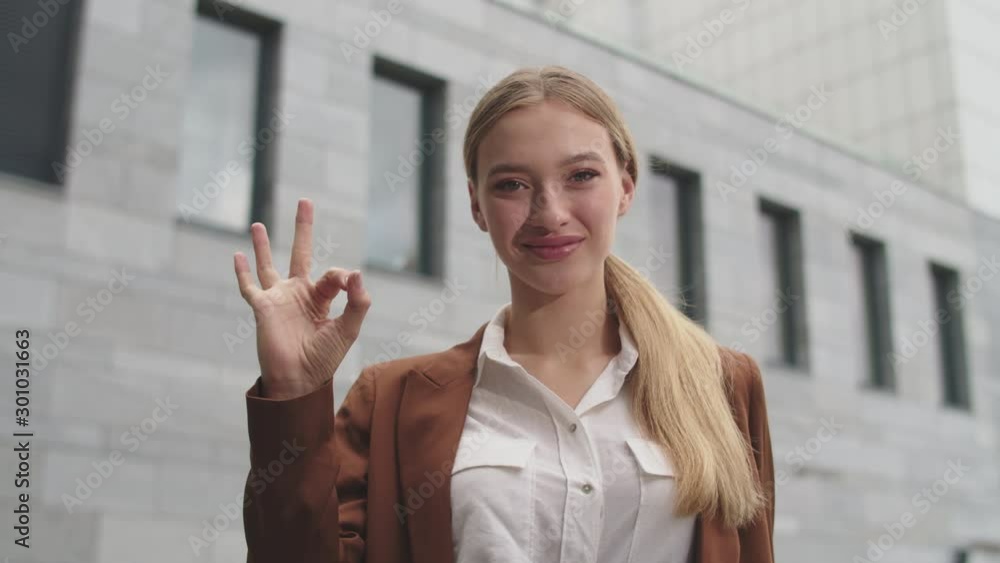 Portrait of glad young lady showing OK hand gesture smiling looking at camera standing against big business building. Portrait of glad young lady showing OK hand gesture smiling looking at camera standing against big business building.