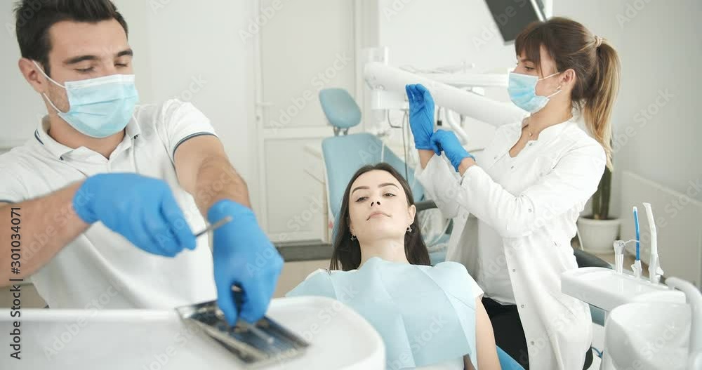 Young male dentist taking tools and checking woman's oral cavity, helpful assistant waiting near, stomatology Young male dentist taking tools and checking woman's oral cavity, helpful assistant waiting near, stomatology