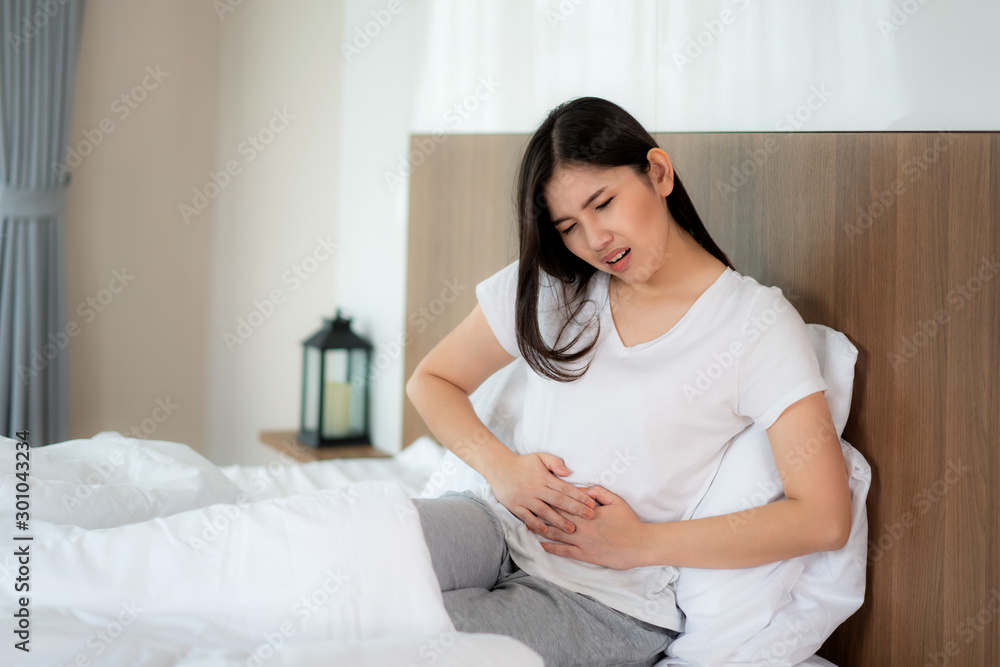 Asian woman unhappy lying on the bed looking sick, suffers from stomach ache in the bedroom, stomachache because of menstruation and eating spoiled food, Chronic gastritis. Abdomen bloating concept Asian woman unhappy lying on the bed looking sick, suffers from stomach ache in the bedroom, stomachache because of menstruation and eating spoiled food, Chronic gastritis. Abdomen bloating concept