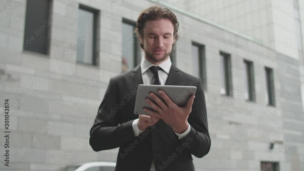 Charming young man in a business suit uses a tablet on the background of a big building. Charming young man in a business suit uses a tablet on the background of a big building.