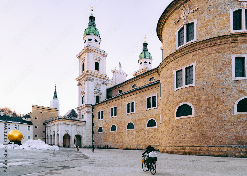 Fototapeta premium Woman on bicycle at Salzburger Dom Salzburg Cathedral in Austria in winter with snow. View of street at Church, old Austrian town. Cityscape of Mozart city, Europe. Architecture of religious building
