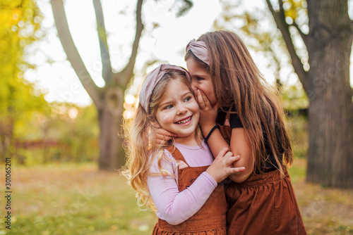 Portrait of two little sisters in a park, whispering secrets.