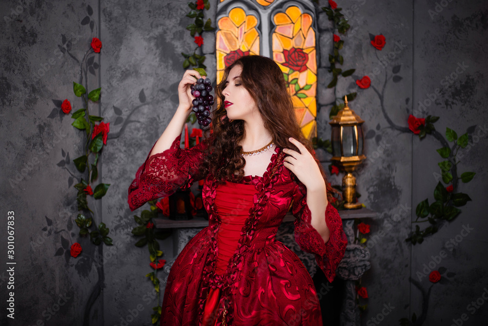 A beautiful girl in a magnificent red dress of the Rococo era stands against a fireplace, a window and flowers with a bunch of grapes in her hands. A beautiful girl in a magnificent red dress of the Rococo era stands against a fireplace, a window and flowers with a bunch of grapes in her hands.