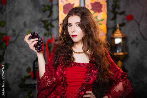 A beautiful girl in a magnificent red dress of the Rococo era stands against a fireplace  a window and flowers with a bunch of grapes in her hands.