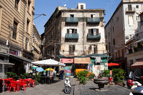 Fototapeta Naklejka Na Ścianę i Meble -  Palermo, Italy, September 19, 2019: Square with a fountain, a parked motorbike, plants and shops around with awnings and facades of buildings in poor condition
