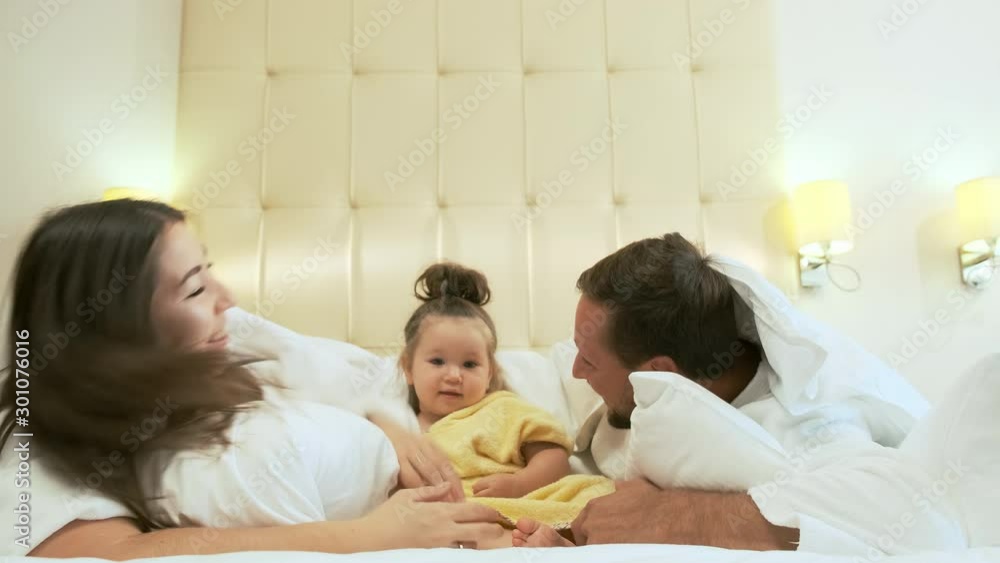 Adorable multiethnic family is playing lying in bed. Mom and Dad have fun with their little daughter