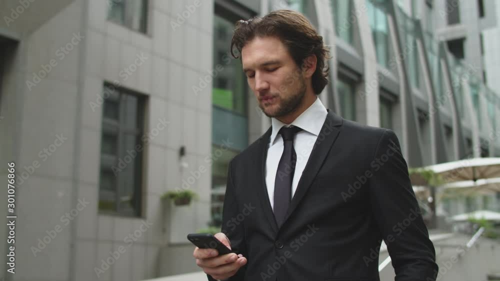 A young guy in a black formal suit is typing a text message on his smartphone in the business district of the city. A young guy in a black formal suit is typing a text message on his smartphone in the business district of the city.