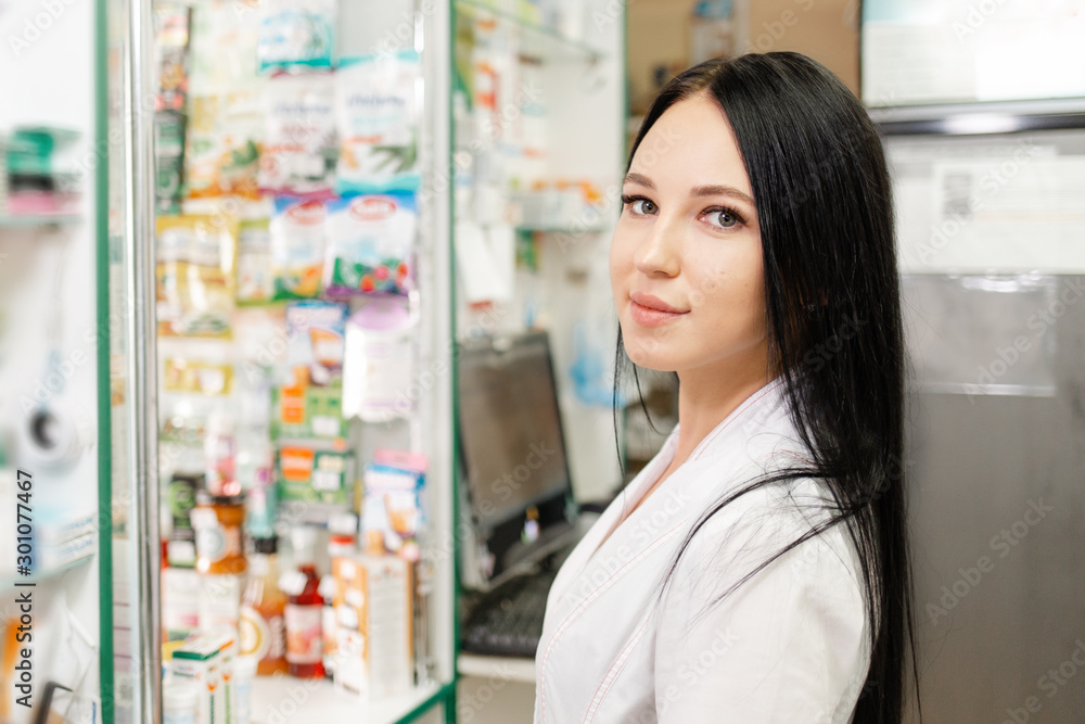 Medicine, pharmacy and optics. Portrait of a woman doctor or pharmacist in a white coat, looking with a smile. In the background a medicine Cabinet and a computer monitor