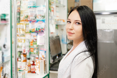 Medicine, pharmacy and optics. Portrait of a woman doctor or pharmacist in a white coat, looking with a smile. In the background a medicine Cabinet and a computer monitor Free Download Medicine, pharmacy and optics. Portrait of a woman doctor or pharmacist in a white coat, looking with a smile. In the background a medicine Cabinet and a computer monitor - GFXLoad