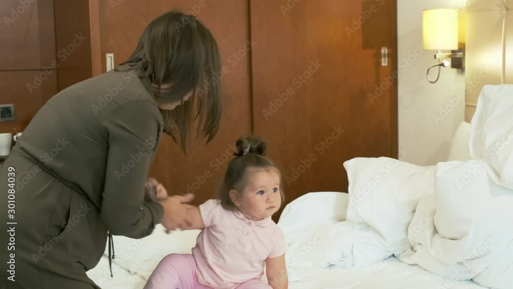 Young Asian mom dresses her little daughter. She is sitting on the bed in a hotel room Young Asian mom dresses her little daughter. She is sitting on the bed in a hotel room