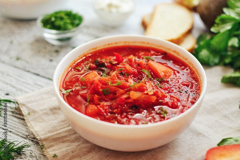 Appetizing Ukrainian and Russian borsch. Tomato soup in a white bowl. Vegan soup with tomato, sweet pepper, cabbage, carrots, beets and parsley. Food on a wooden background with textiles. Appetizing Ukrainian and Russian borsch. Tomato soup in a white bowl. Vegan soup with tomato, sweet pepper, cabbage, carrots, beets and parsley. Food on a wooden background with textiles.