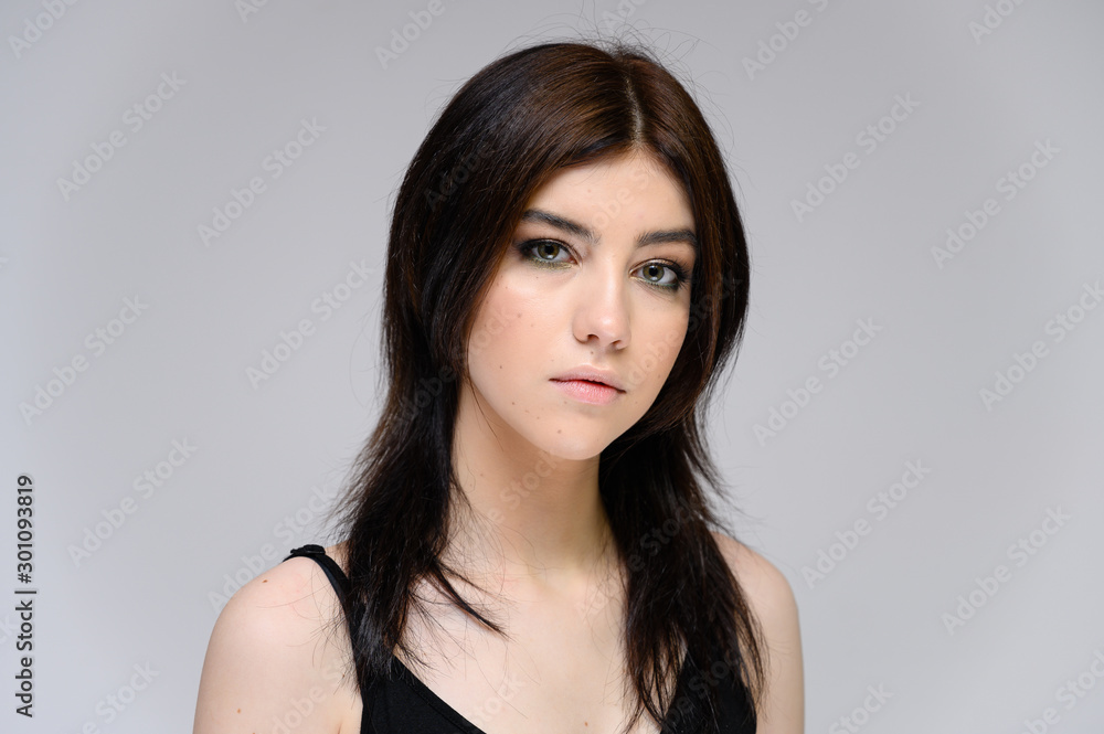 Fototapeta premium Close-up portrait of a young pretty girl with long black hair, clean skin on a gray background. The concept of cosmetics for the face, great makeup, fashionable hairstyle.