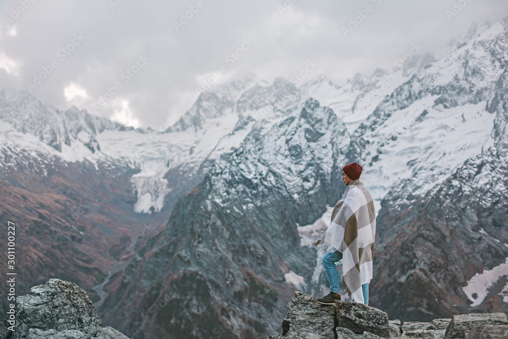 Young guy wrapped in plaid blanket standing on mountain peak