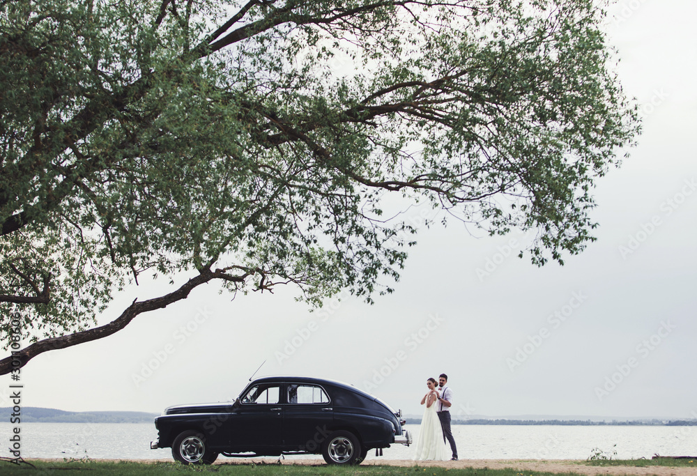 bride and groom standing near a retro car on the background of the lake
