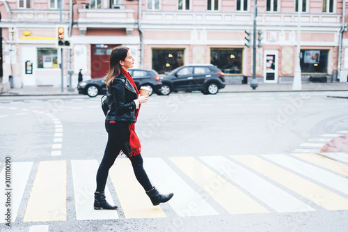 A beautiful brunette woman crosses the road on pedestrian crossing in a black leather jacket and a red scarf and hold in hand a hot drink.