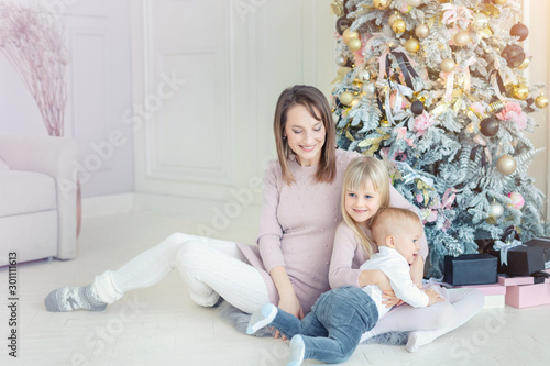 Portrait of cute adorable little blond children with mother enjoy sitting near christmas tree in cozy living room at home . Happy mom with little son and daughter. New year holidays happy family © Kirill Gorlov