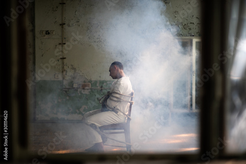 Принти на полотні A crazy man in a straitjacket is tied to a chair in an abandoned old clinic