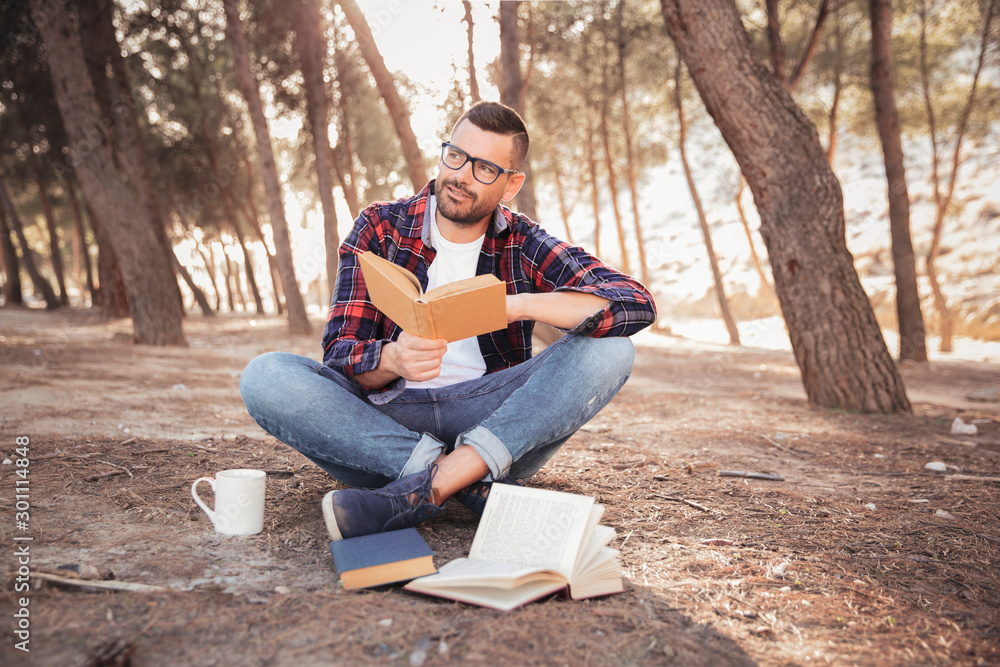 Fototapeta premium Hombre joven con lentes y camisa de cuadros, sentado en el suelo en medio del bosque, leyendo sonriente un libro al aire libre