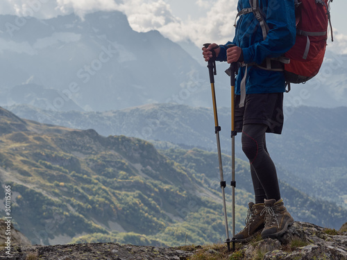 Young hiker standing on cliff edge; legs view; brave man with climbing equipment, backpack, trekking poles enjoying a freedom, looking for route; mountain ranges, cloudy sky on background