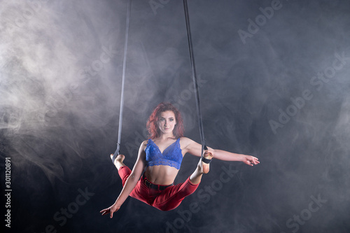 Female athletic, sexy and flexible aerial circus artist with redhead on aerial straps on black background