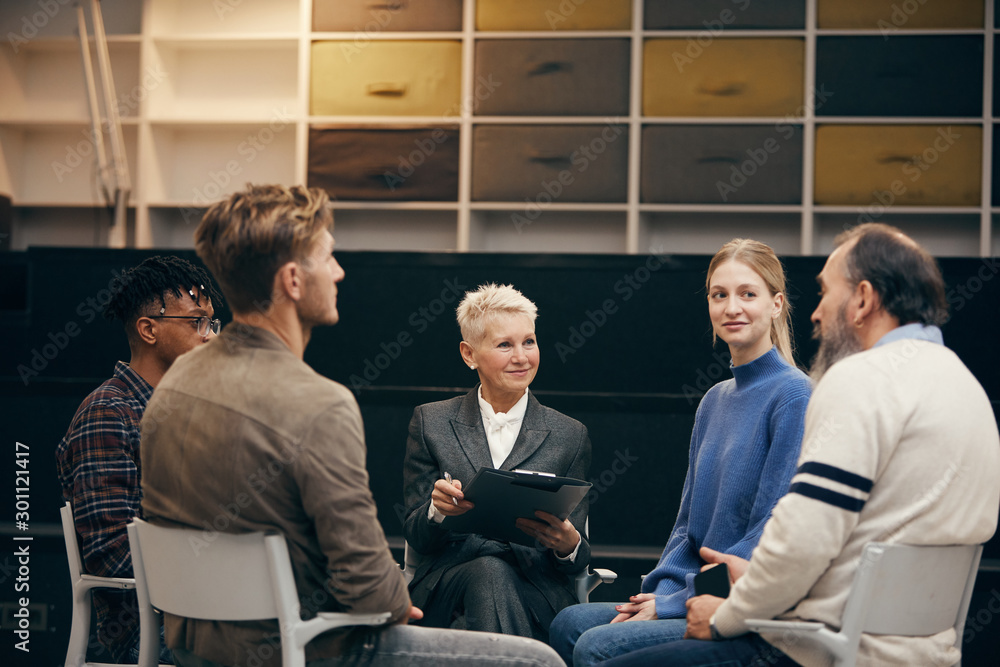 Mature woman with short blond hair making notes while people telling their problems during therapy Mature woman with short blond hair making notes while people telling their problems during therapy