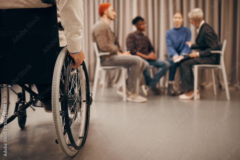 Close-up of disabled man sitting on wheelchair with group of people talking to each other in the background Close-up of disabled man sitting on wheelchair with group of people talking to each other in the background