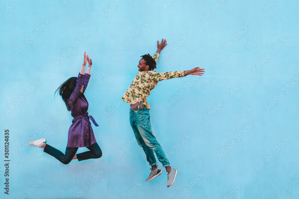 Fototapeta premium African couple jumping outdoor with blue wall in background - Crazy happy latin people having fun together - Youth, friendship and fashion concept - Main focus on man face