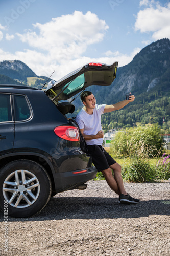 Young handsome traveler man take selfie on phone while sitting in car trunk neat perfect mountains background. © F8  \ Suport Ukraine