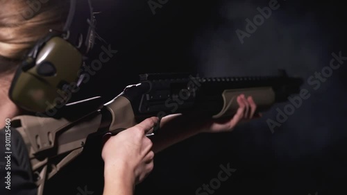 Woman aiming and firing shotgun with recoil, wearing ear protection in shooting range with dark background. photo