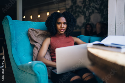 Portrait of intelligent manager of cafeteria with loft interior resting in comfortable chair with modern laptop computer for planning week via organizer app, skilled female student with netbook
