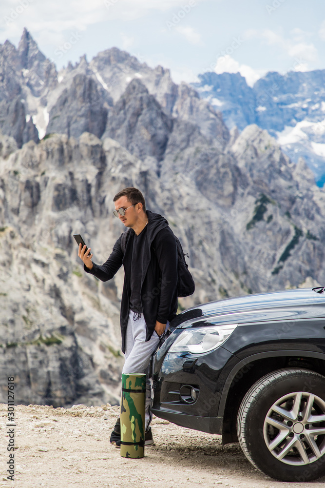 Young handsome man sitting on hired car and talking on the mobile phone on mountains background. Car hire. Car trip.