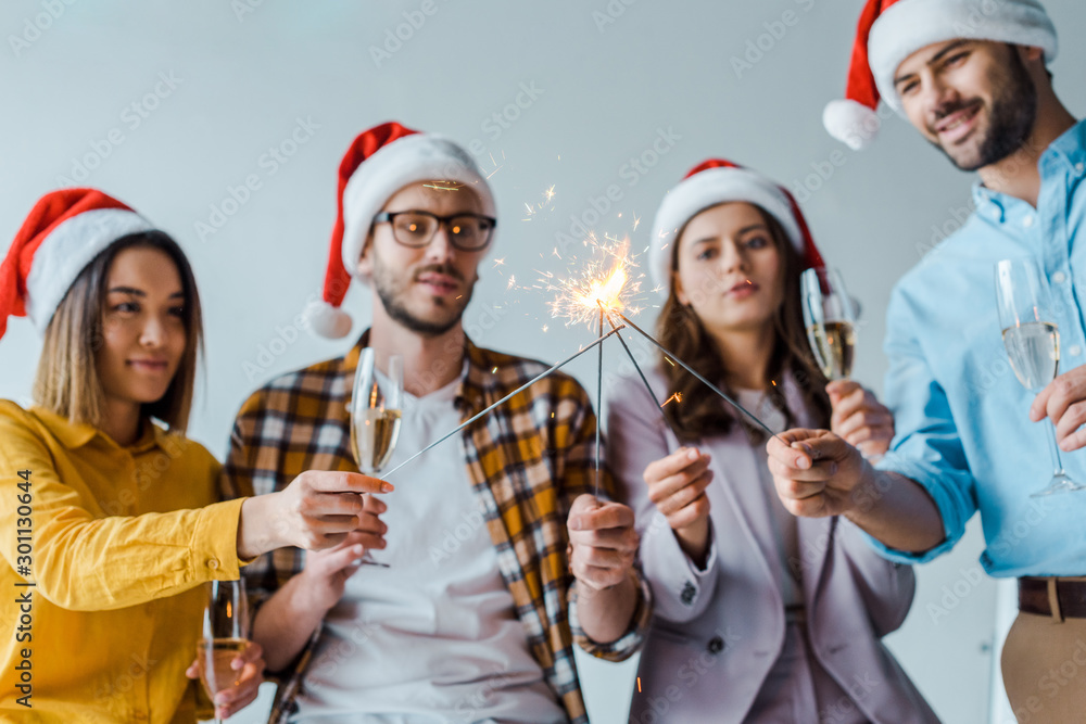 Fototapeta premium selective focus of cheerful businessmen and multicultural businesswomen holding sparklers and champagne glasses in office