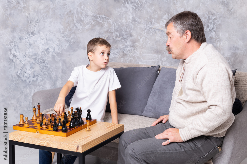 Fotka „Grandfather playing chess with grandson indoor. The boy and his  grandpa are sitting on the sofa in the living room and talking together.  Senior man teaches a child to play chess“ ze
