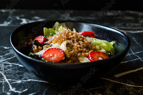 Buckwheat Salad with Strawberry Slices and Green Leaves in Black Bowl on Dark Granite Surface.