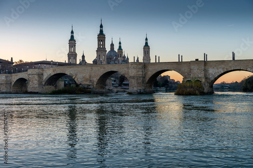 View of the medieval stone bridge in front of the Cathedral of El Pilar, Zaragoza (Spain), on the banks of the River Ebro, during the sunset.