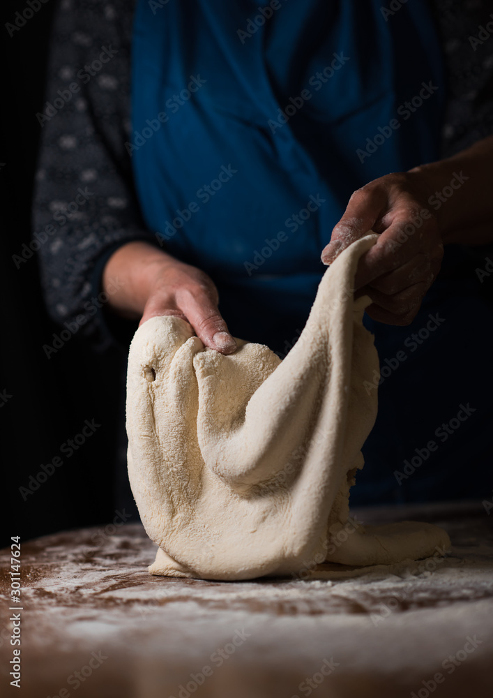 The wrinkled hands of a woman hold a rendered dough on a dark background.