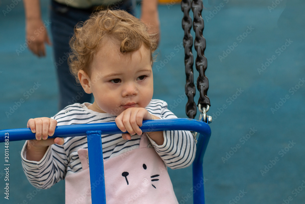 The little girl rides on a swing. Caucasian baby kid with curls on the playground The little girl rides on a swing. Caucasian baby kid with curls on the playground