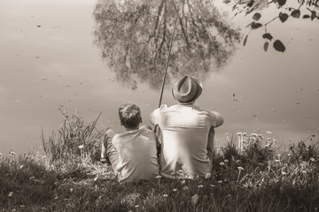 Back view of happy family on summer vacations concept. Father and son fishing together at river bank at scenic landscape background of fresh green grass and blue calm water. Horizontal sepia photo.