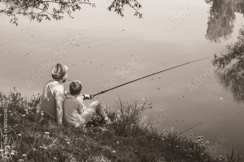 Back view of happy family on summer vacations concept. Father and son fishing together at river bank at scenic landscape background of fresh green grass and blue calm water. Horizontal sepia photo.