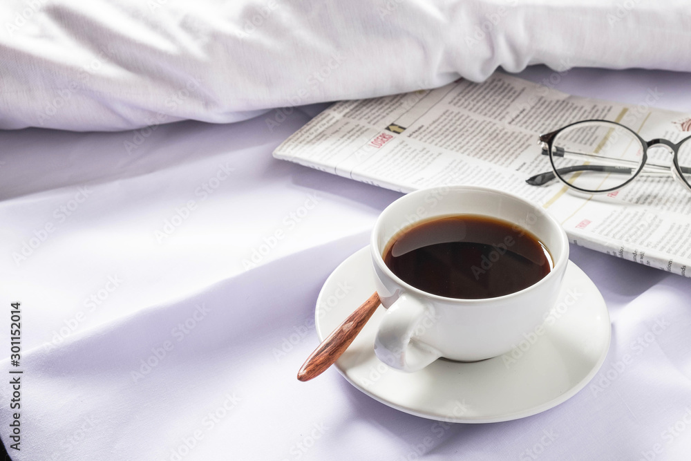 A cup of coffee and a newspaper on a white bed in the morning. - obrazy, fototapety, plakaty 