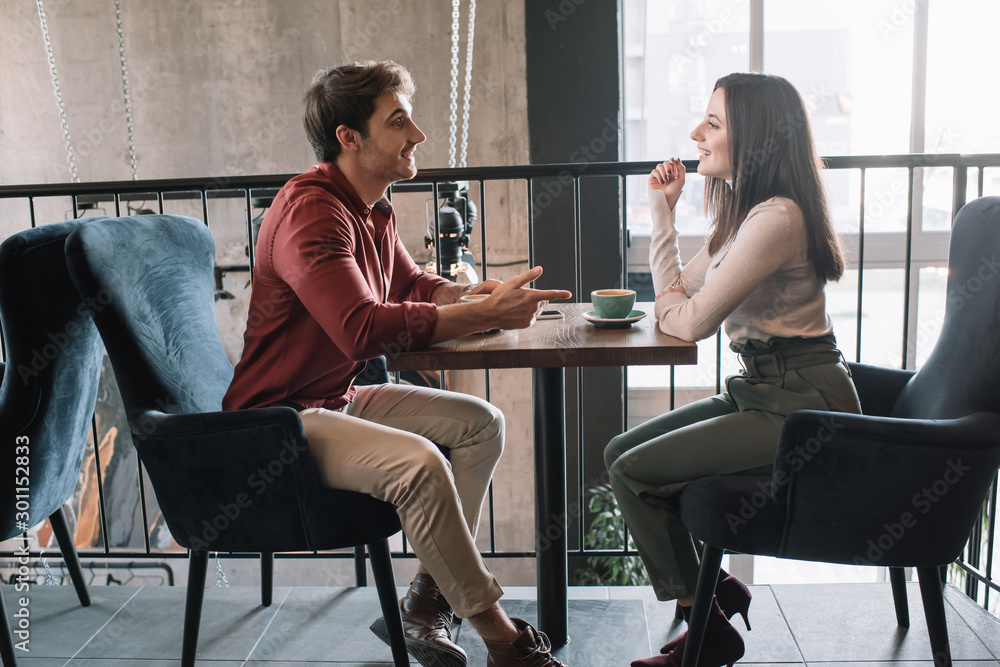 side view of cheerful couple talking while drinking coffee on balcony in coffee shop