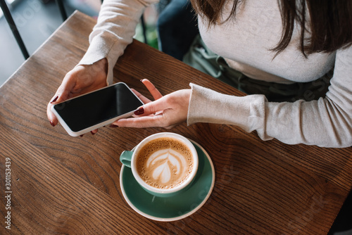 Fotografie cropped view of young woman sitting at wooden table with cappuccino and holding
