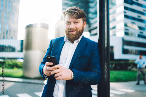 Half length portrait of cheerful successful man in formal wear holding cellphone gadegt in hands and smiling at camera during time in financial district, positive bearded boss with smartphone photo