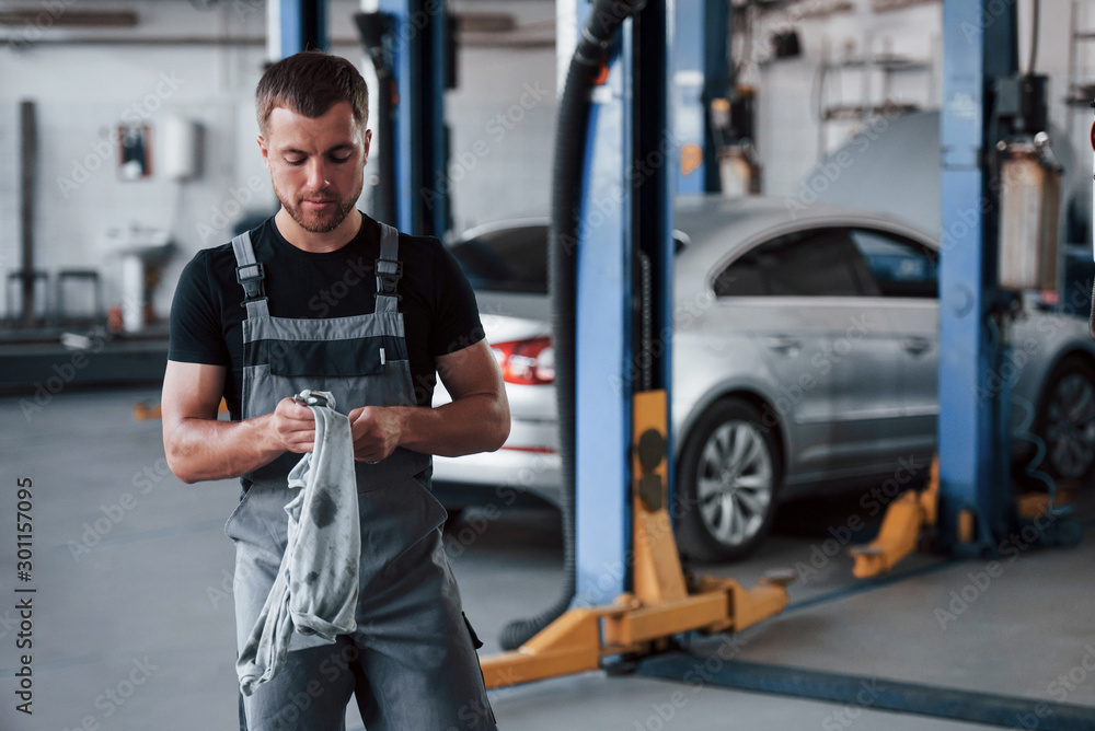 Order is done well. Man in black shirt and grey uniform stands in garage after repairing broken car Order is done well. Man in black shirt and grey uniform stands in garage after repairing broken car